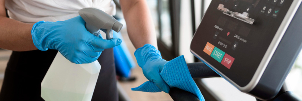 Reinigungskraft mit blauen Handschuhen reinigt ein Display am Fitnessgerät – gründliche Desinfektion im Fitnessstudio