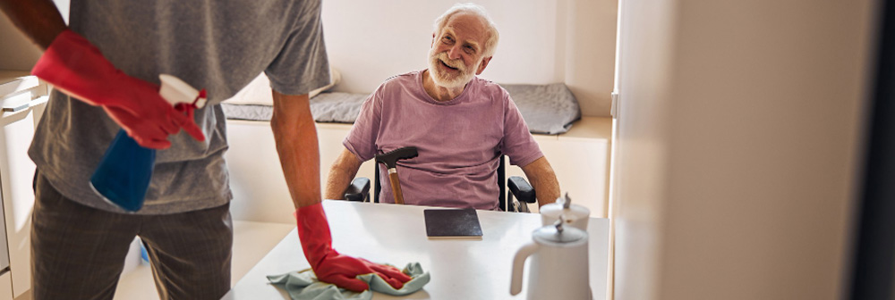 Reinigungskraft wischt Tisch in Pflegezimmer, älterer Mann im Rollstuhl lächelt – sanfte Reinigung im Seniorenheim